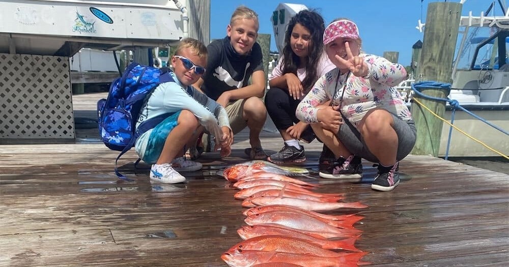Full Day Fishing Charter Image 1 Four kids crouching on a dock beside a row of freshly caught fish.