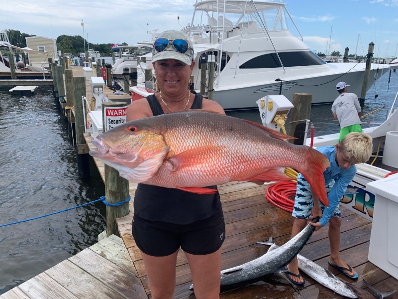 Person holding a large fish on a dock, with boats and water in the background.