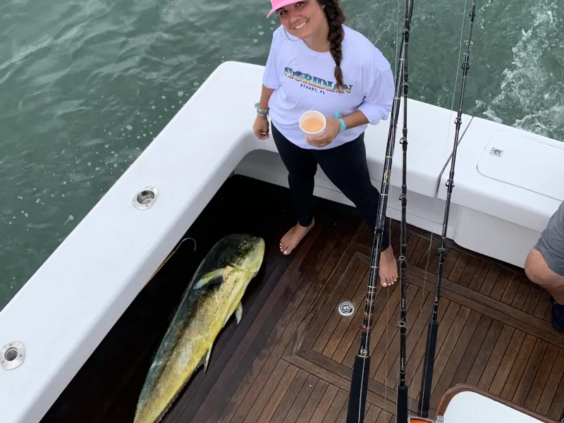 Woman on boat deck smiles near caught large fish, holding a drink.
