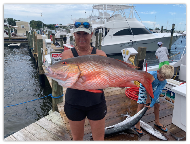 Person holding a large fish on a dock, with boats in the background.