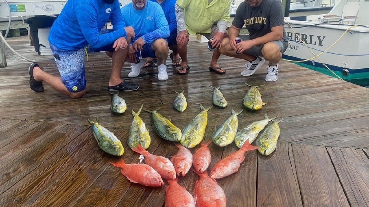 Five men posing on a dock with a display of caught fish, including red and yellow species.