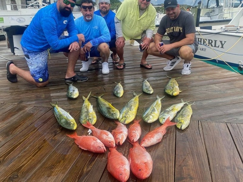 Five men posing on a dock with a display of caught fish, including red and yellow species.