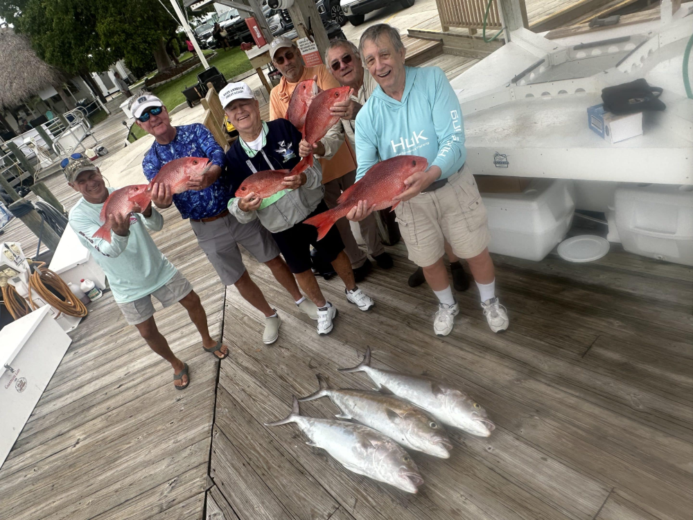 Six people hold red fish on a dock with three large silver fish on the ground.