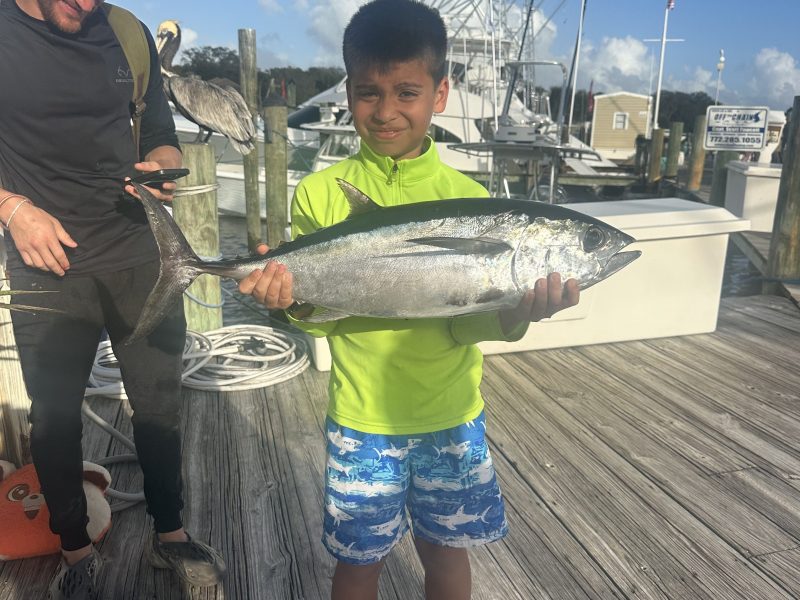 Boy in bright shirt holding large fish on dock; man smiling beside him.