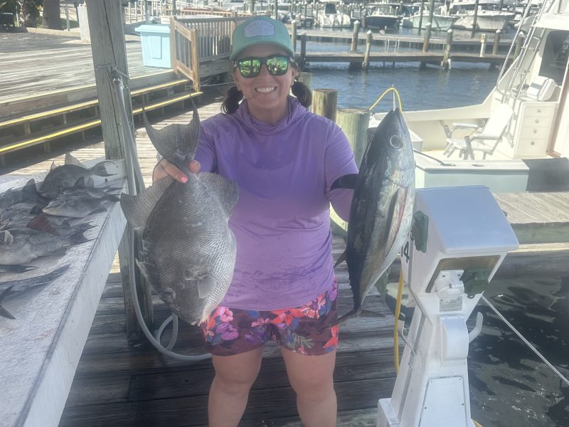 Person holding two fish on a dock with boats in the background.